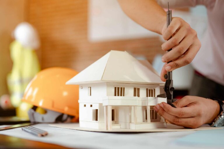 close up of male architect hands measuring and making model house on the desk at sunset 768x511